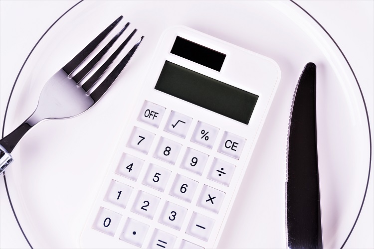 A calculator sitting next to a fork and knife on a white plate, representing the comparison of calorie counting and healthy eating for weight loss